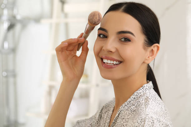 Beautiful young woman applying face powder with brush in bathroom at home, make up essentials